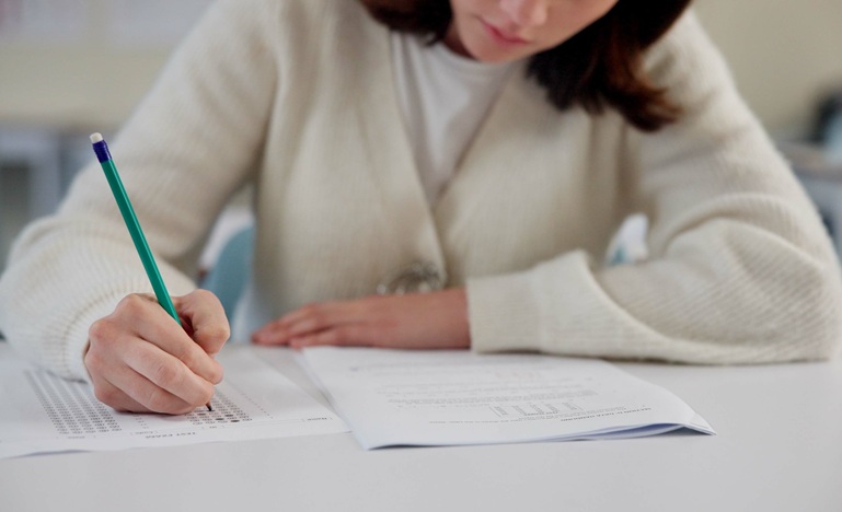 Young woman seated at long table filling out multiple-choice exam in booklet, using pencil and looking downward at page