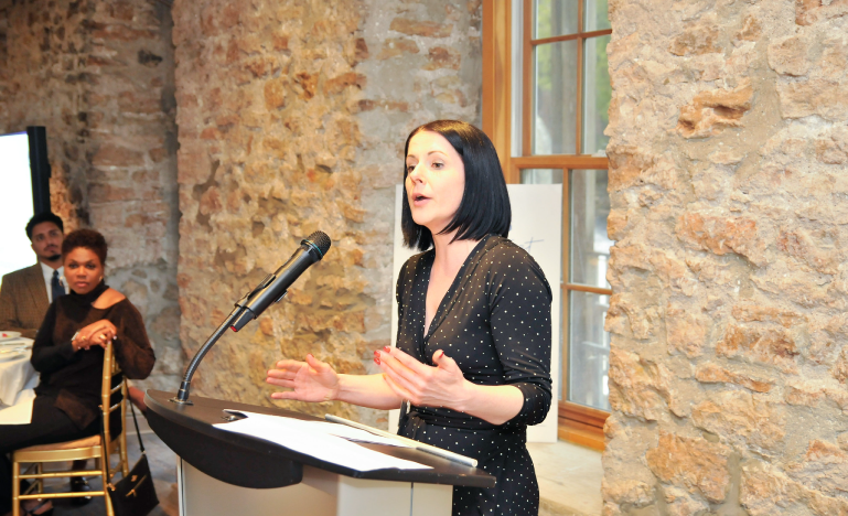Kelly McDermott, wearing a black polka dot dress, speaking at a podium in a rustic venue with exposed stone walls, while attendees listen.