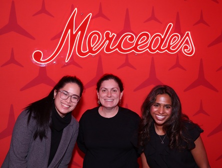 Author Hanaa poses next to her supervisor and a colleague against a red wall featuring the Mercedes-Benz logo and the word Mercedes written above their heads