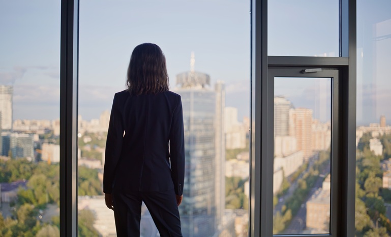Woman in a business suit stands with her back to the camera, looking out the floor-to-ceiling window of an office at the skyscrapers and buildings and open sky around her