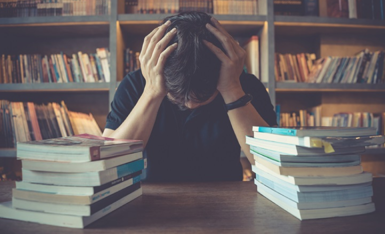 Stressed student sitting at a table with stacks of books, holding their head in frustration, in a library setting.