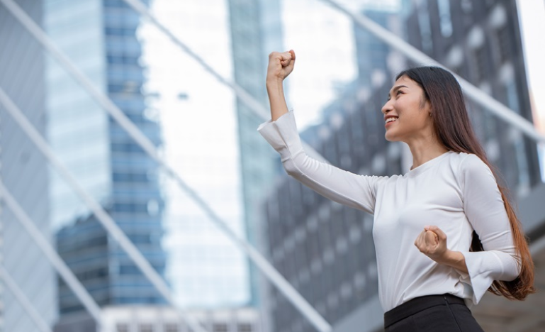 Confident woman in a white blouse celebrating success with a fist pump in front of modern glass buildings.