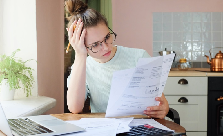 Worried woman with glasses reviewing bills at a kitchen table with a laptop and calculator.