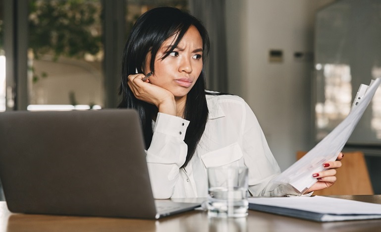 Woman seated at desk with laptop, viewing printed document with confusion