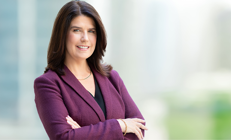 Photo of OBA President Kathryn Manning, standing against city and parkscape backdrop, wearing purple jacket, with arms folded loosely across chest