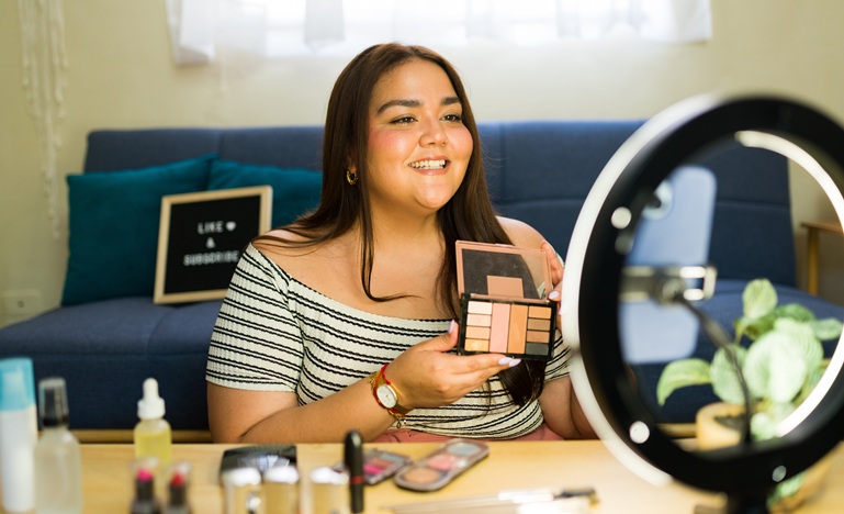 a young woman is seated at vanity table holding an open palette of eye shadow and staring into a camera with a ring light around it as she talks about the product