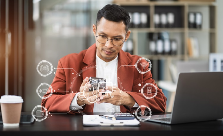Man in glasses and a brown blazer analyzing a small device with digital technology icons overlaid, sitting at a desk with a laptop and coffee cup.