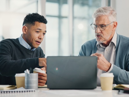 A young male lawyer sits next to an older male client at a table explaining information that appears on the laptop screen in front of them
