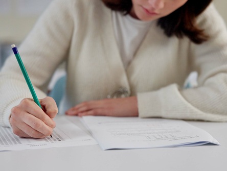 Young woman seated at long table filling out multiple-choice exam in booklet, using pencil and looking downward at page