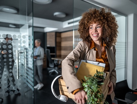 A young woman carrying a box of belongings, including a plant and headphones, enters a new workplace and looks around
