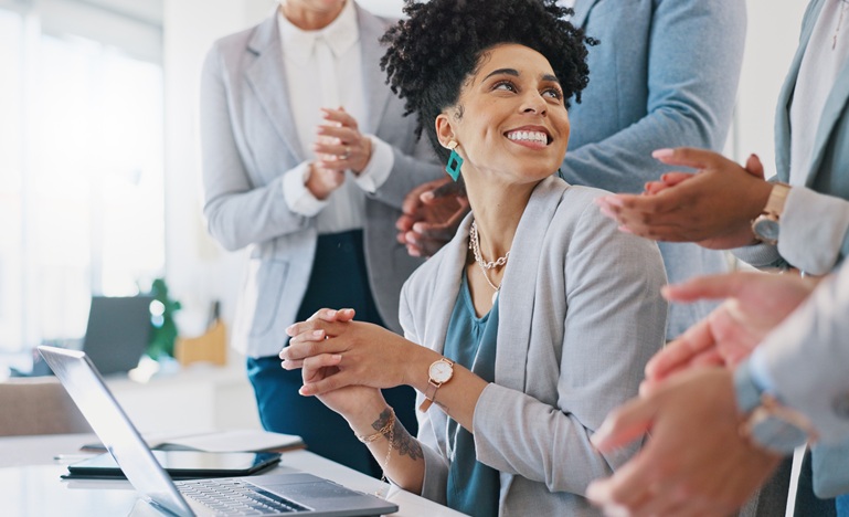 a women seated in front of computer at desk is surprised and delighted to be surrounded by coworkers who are applauding her work
