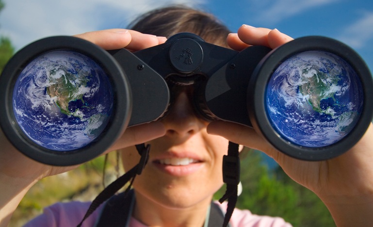 women standing outside in nature holds binoculars up to her eyes and we see reflection of globe in both lenses