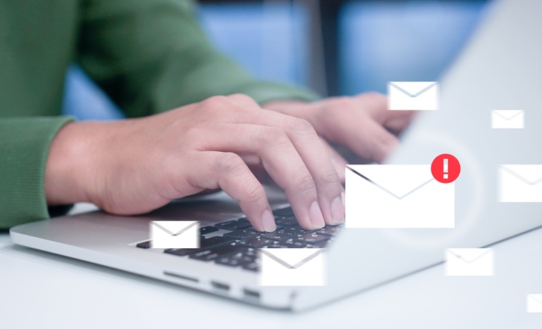 hands type at computer keyboard with email 'envelopes' floating above, one of which is superimposed with a red exclamation mark