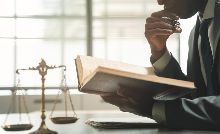 a man seated at a desk with a hand beneath his chin studies a legal book that is open before him next to table-top scales of justice