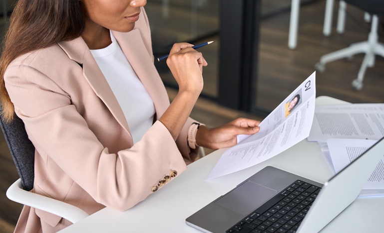 Business woman in salmon-coloured jacket is seated at desk in front of laptop reviewing a resume, with a pen in her hand